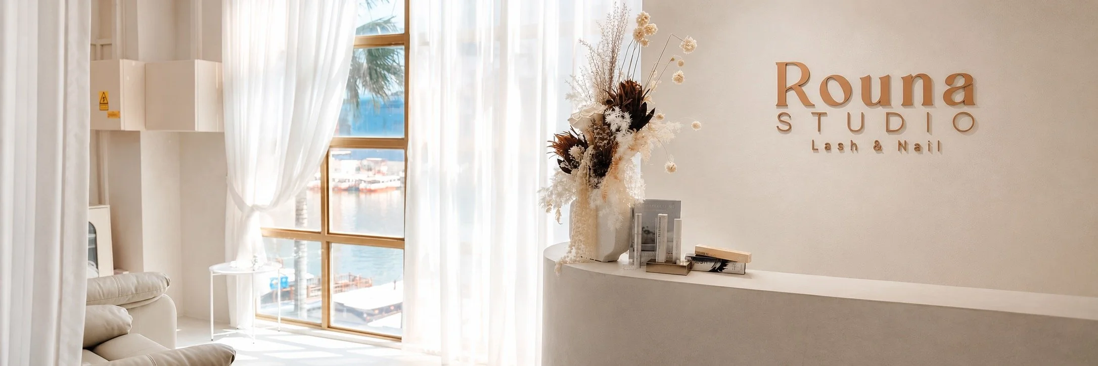 Interior of a beauty studio with white curtains, a large window, and a sign reading 'Rouna Studio Lash & Nail' on the wall. There is a flower arrangement on the reception desk.