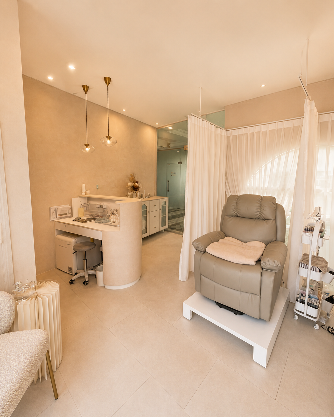 A modern, softly lit room with a beige armchair, a white privacy curtain, and a small white table with medical supplies. There is a desk with a stool, a cabinet with flowers, and pendant lights hanging from the ceiling.
