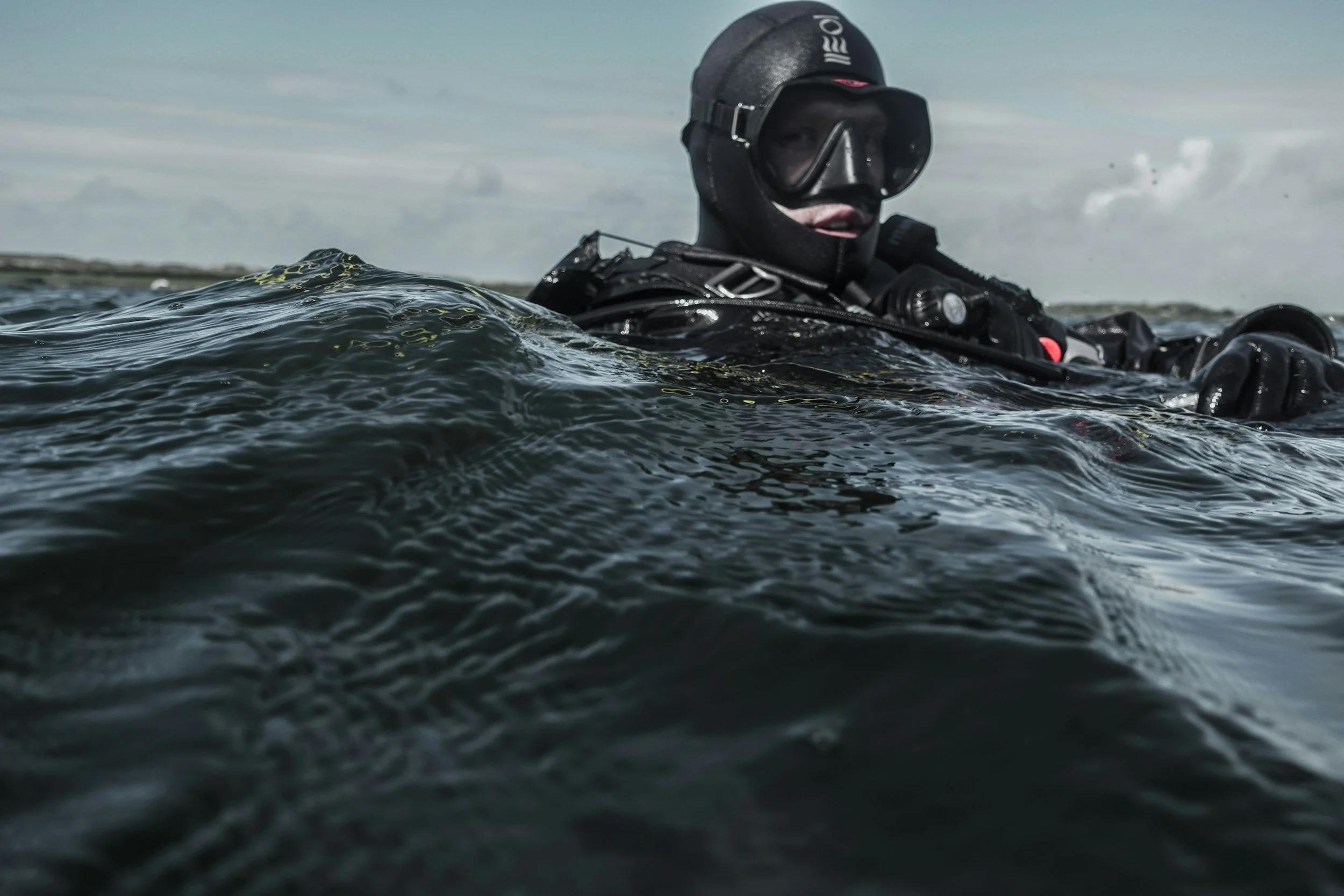 A scuba diver in a black wetsuit and helmet swimming in open water, with waves visible in the foreground and a cloudy sky in the background.