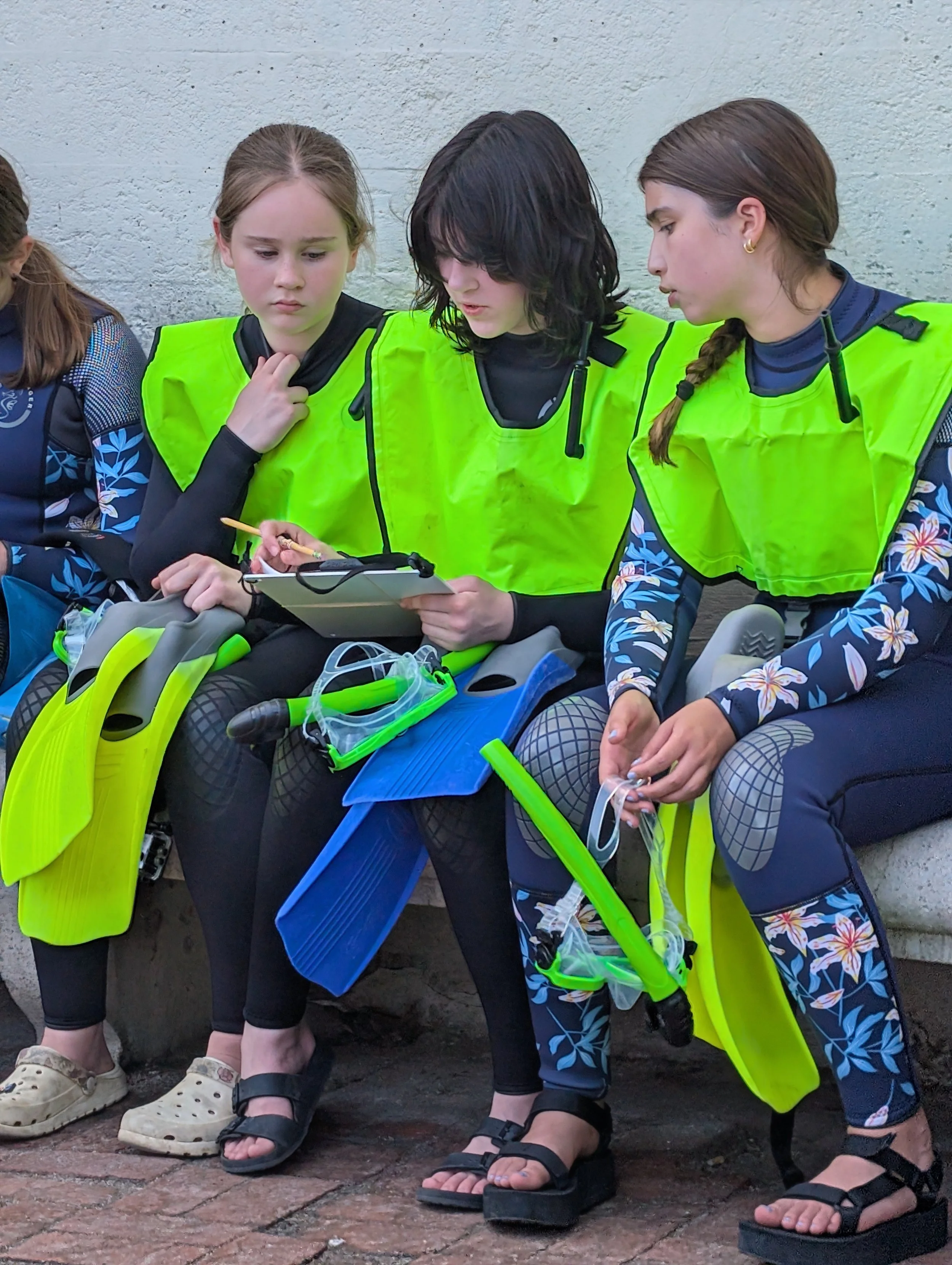 Four young women sitting against a wall, dressed in cyclist gear, with bright yellow-green vests. They are looking at a smartphone held by one of the women. The women are wearing talking goggles around their necks, and their footwear includes sandals and sneakers.