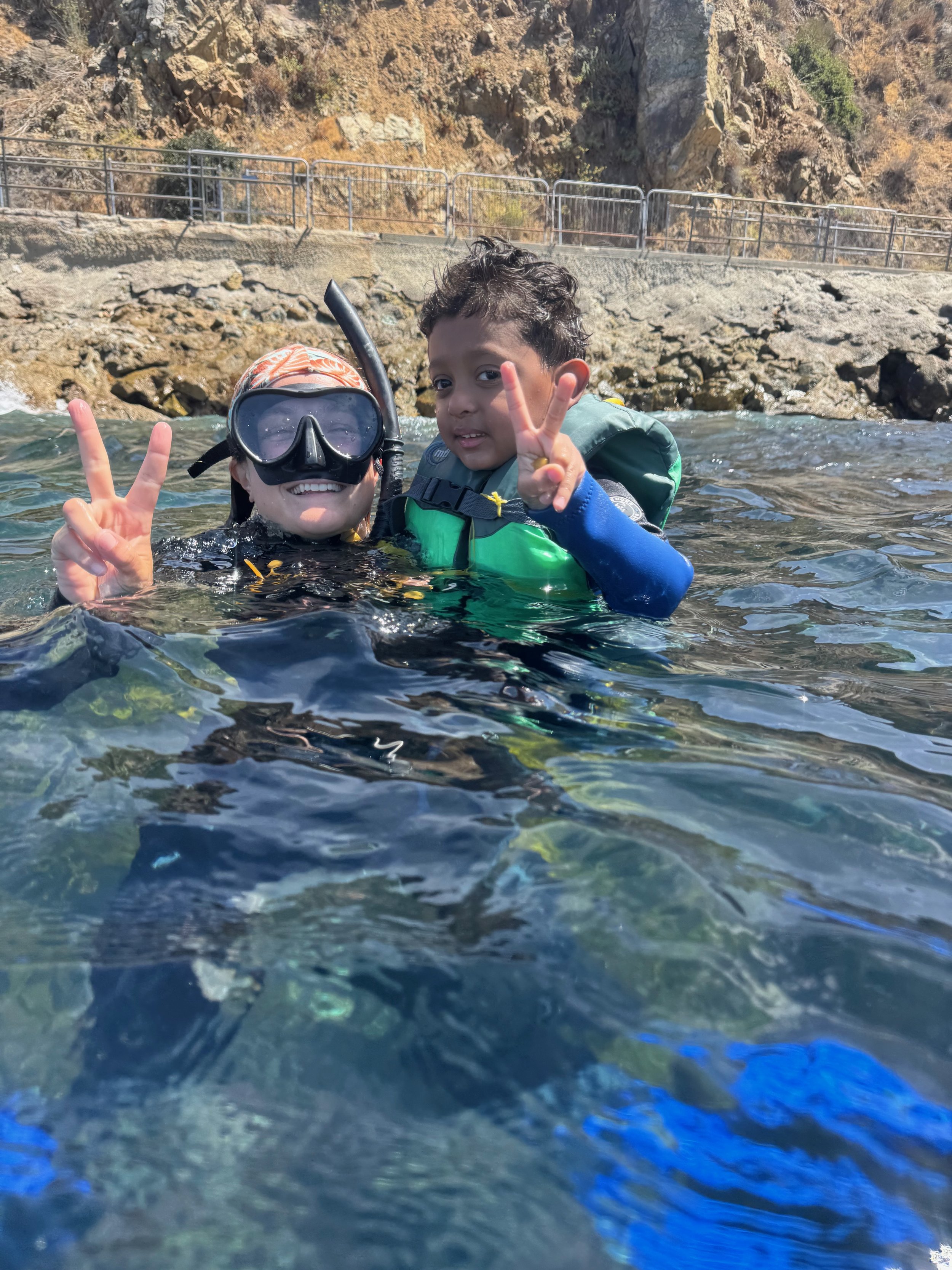 Two children swimming in a river or lake near rocky terrain and a hillside. One child is wearing a snorkeling mask and snorkel, the other is making a peace sign with fingers.