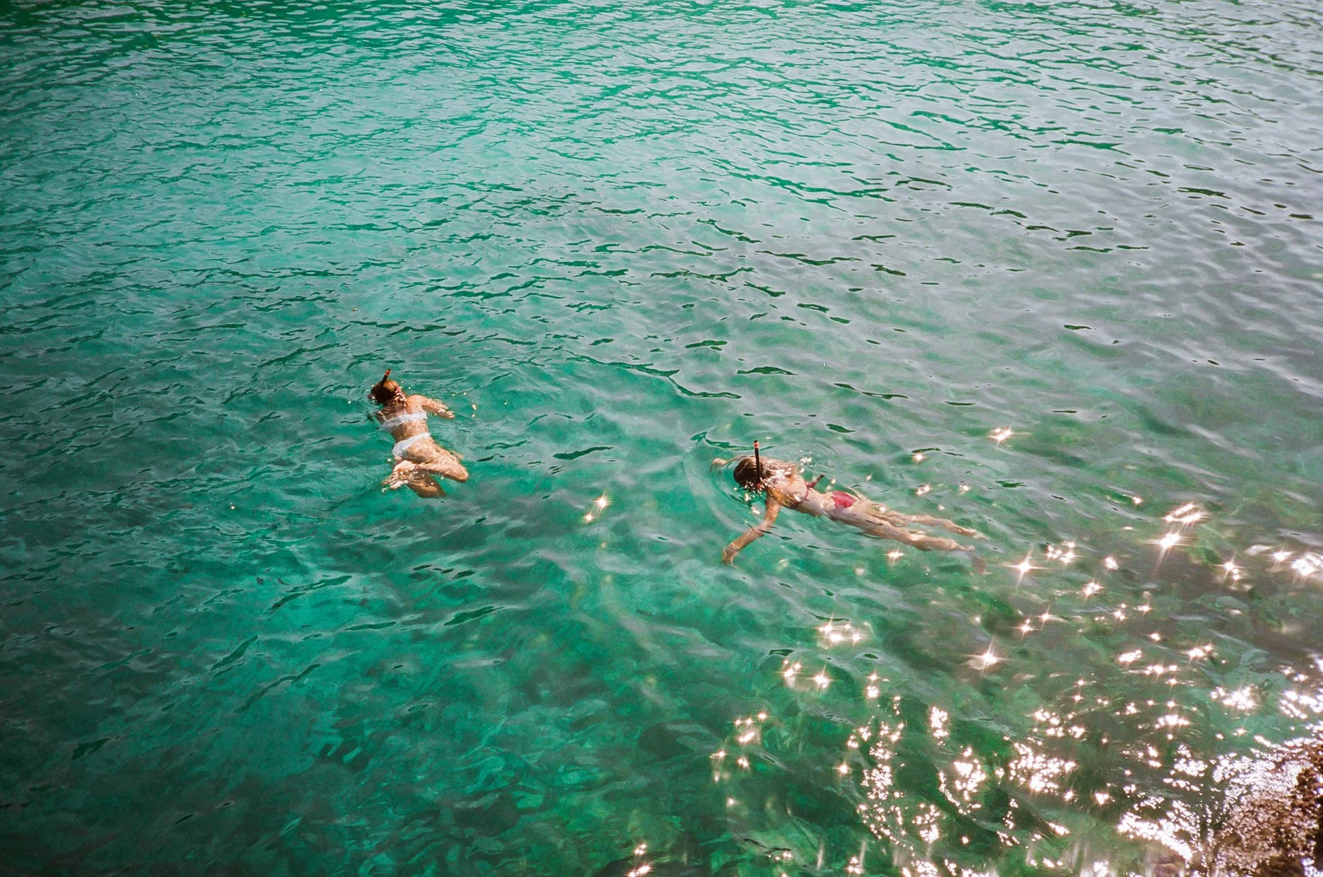 Two children swimming in ocean water, wearing snorkeling gear with fins, near the shore with sunlight reflecting on the water surface.