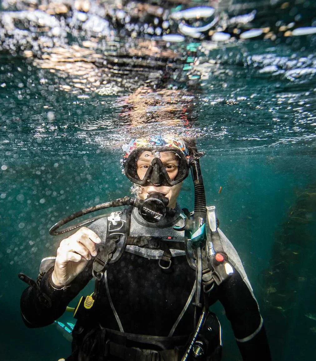 A person wearing scuba diving gear underwater, looking at the camera, with water and bubbles around them.