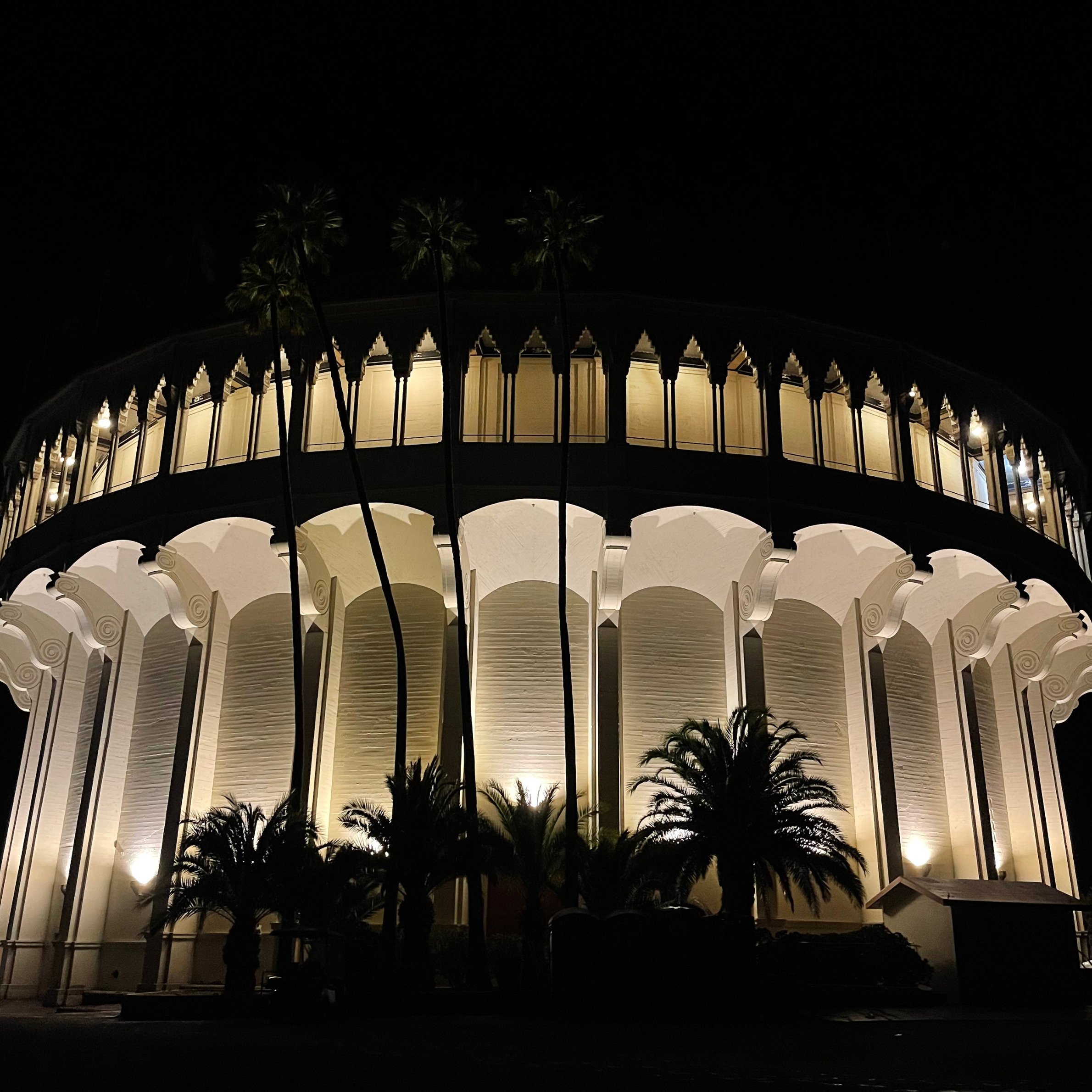 Night view of a brightly lit, circular architectural structure with tall, evenly spaced columns and palm trees in front.