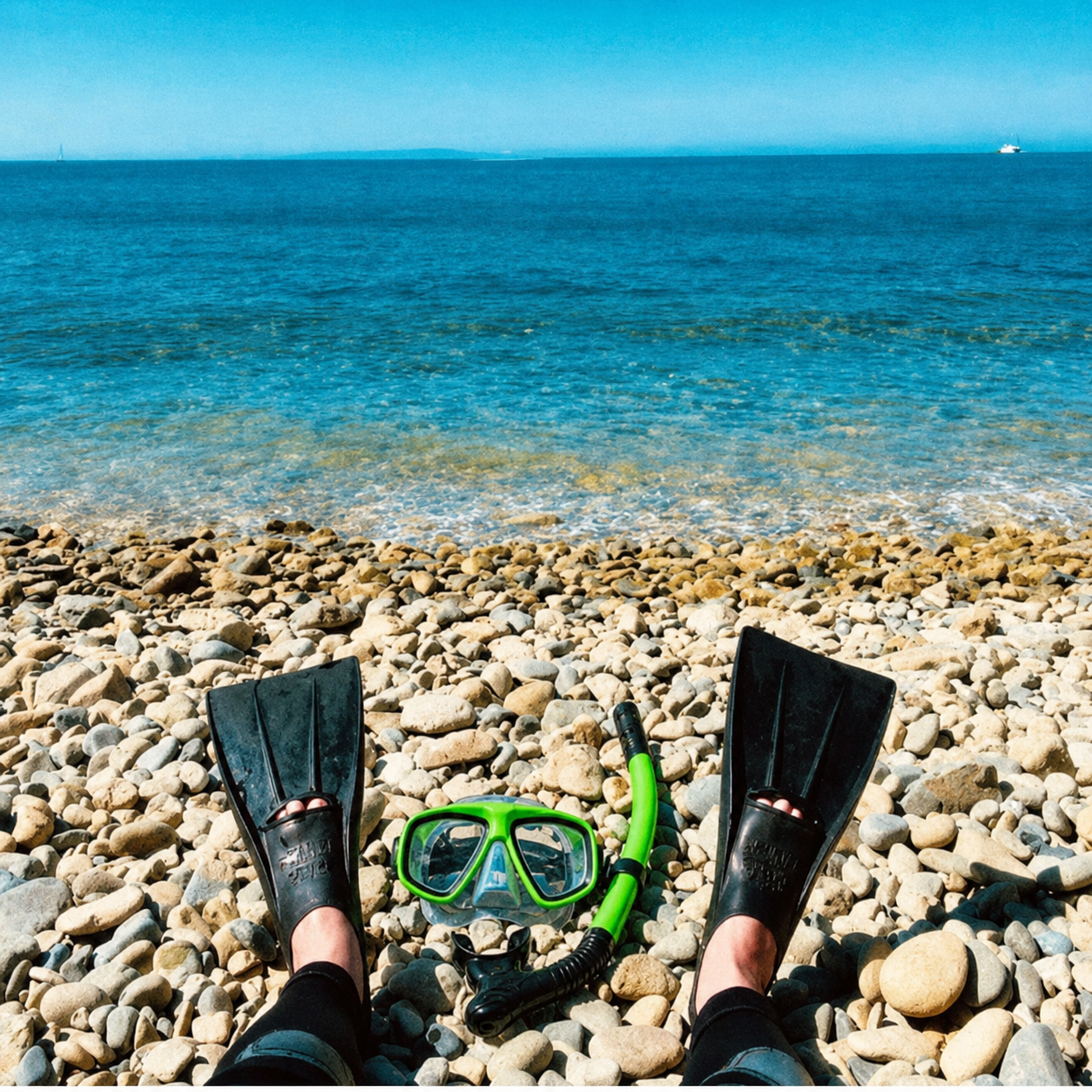 Person relaxing on a pebble beach with snorkeling gear, looking out at the calm ocean and distant ships under a clear blue sky.