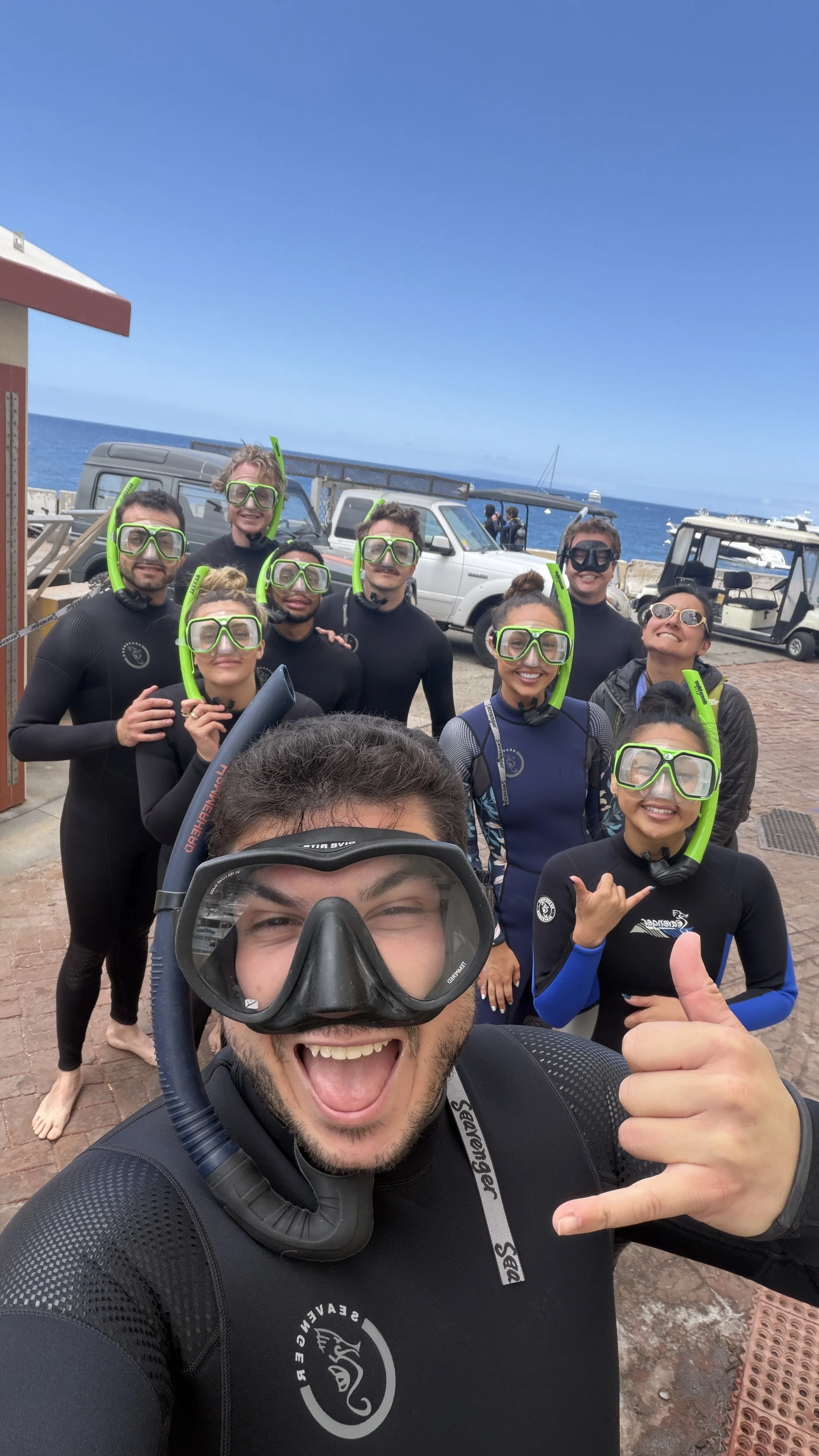 Group of people in wetsuits and snorkeling gear posing for a photo by the ocean, with boats and a clear sky in the background.