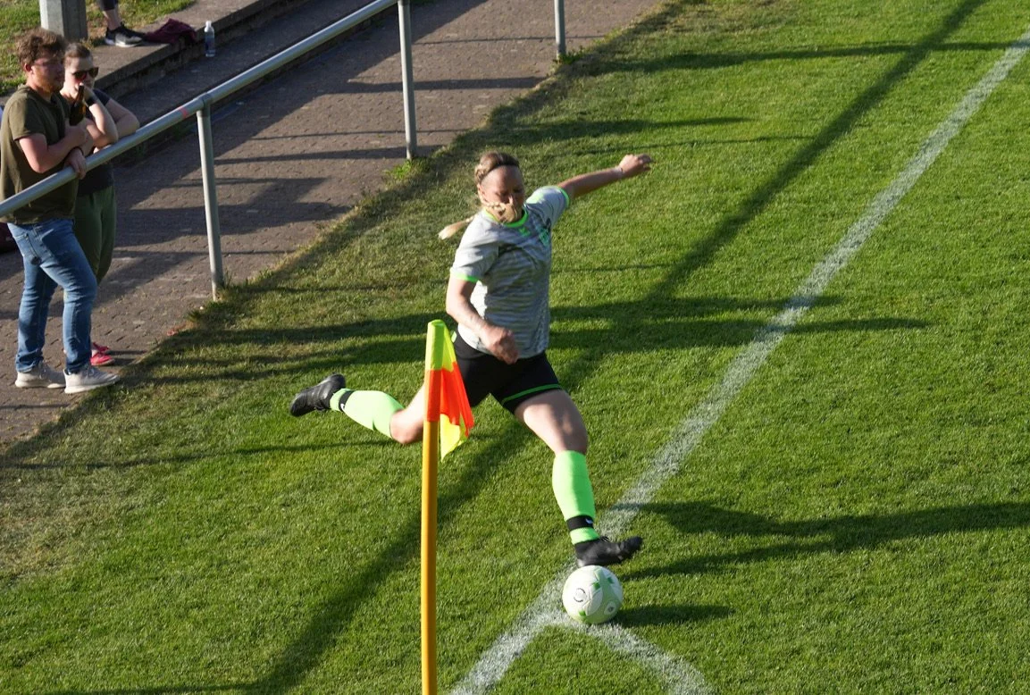 Female soccer player taking a corner kick during a match, with spectators standing behind the railing watching.