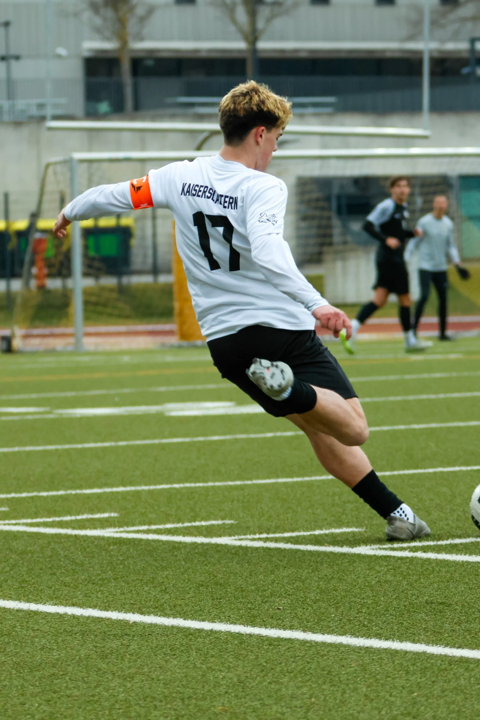 A soccer player wearing a white jersey with the name 'KAISERSLATERN' and the number 17 is kicking a soccer ball on a green field. Two other people, possibly officials or coaches, are in the background near the sideline.