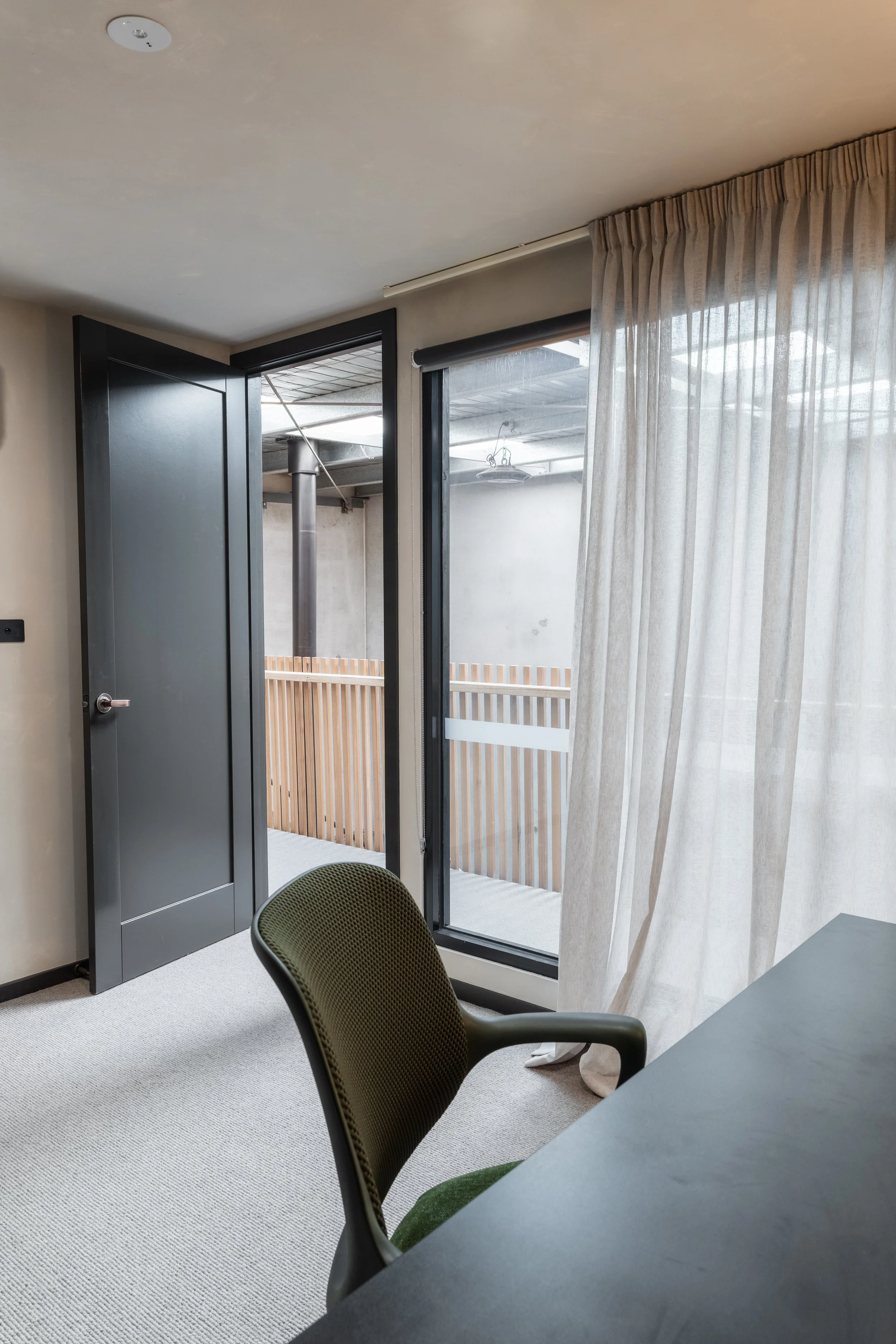 Interior of a room with an open door leading to a small indoor balcony, beige curtains, a black table, and a green office chair with a mesh backrest.