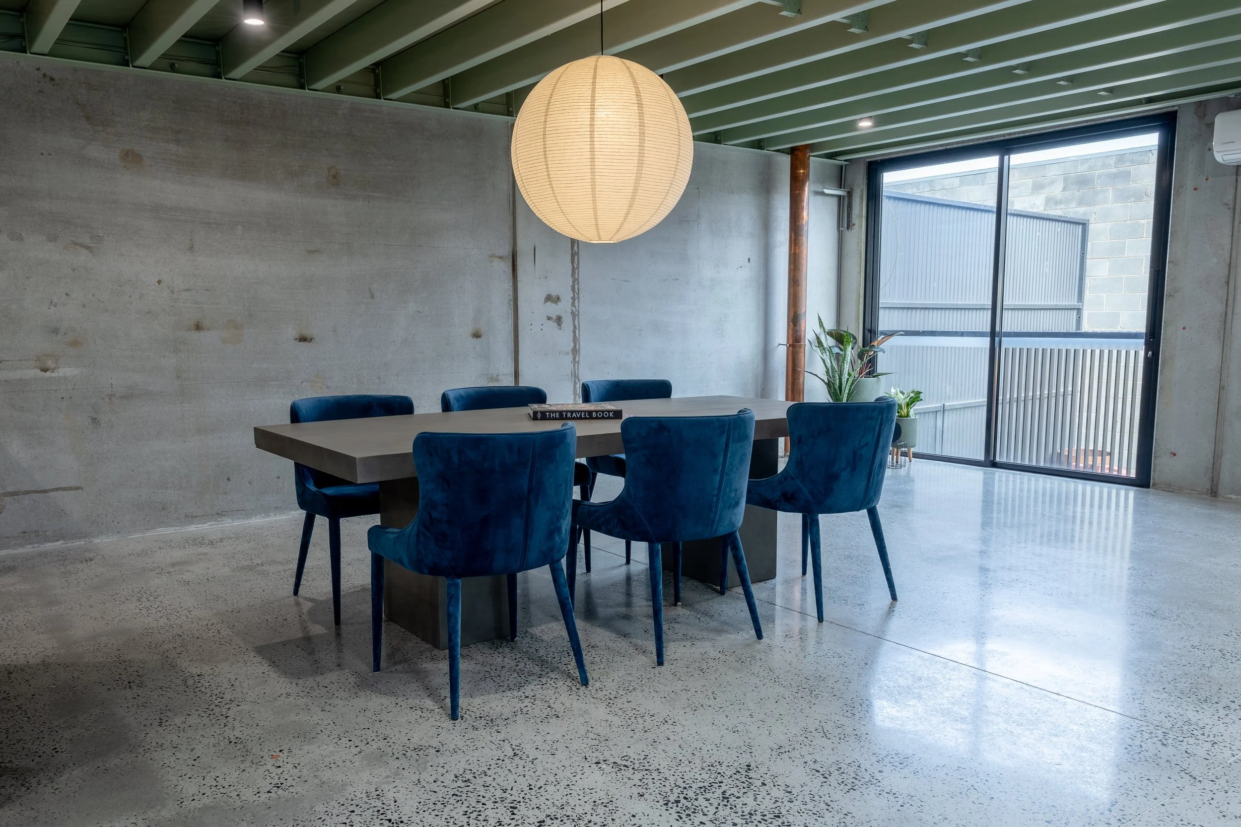Modern dining area with a concrete wall, blue velvet chairs around a rectangular table, large paper lantern hanging from the ceiling, potted plants near a large sliding glass door, and a green painted ceiling with exposed beams.