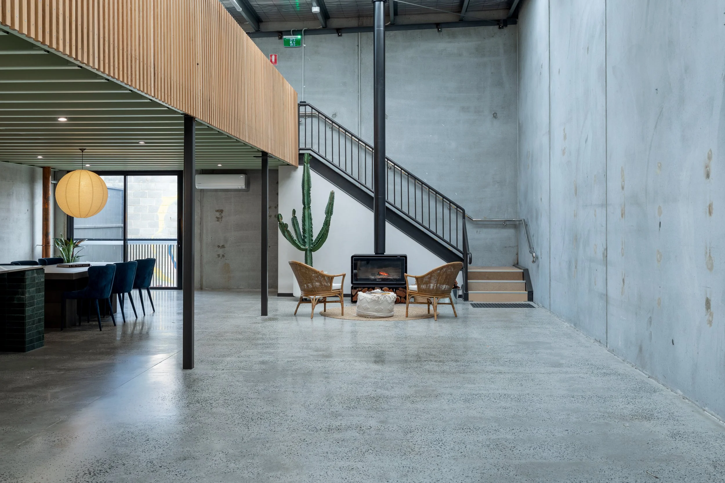 Interior of a modern building with concrete walls, a staircase with black railing, a cactus plant, a seating area with wicker chairs, a fireplace, and large windows.