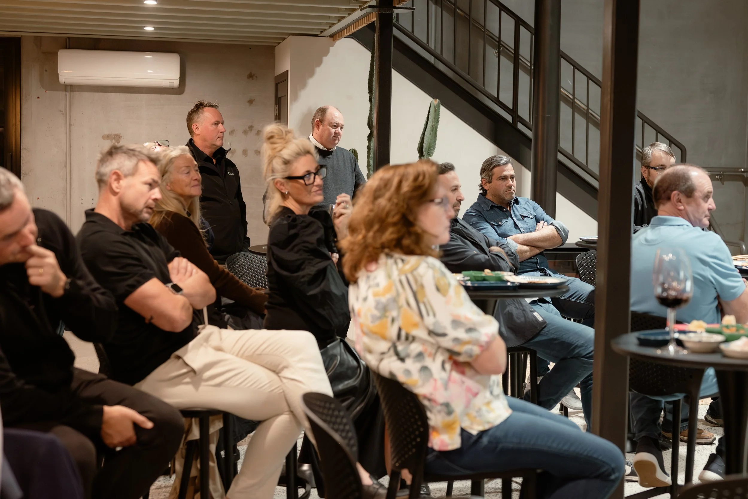 People seated and standing at event, some listening attentively, in urban indoor venue with staircase and cactus plants.