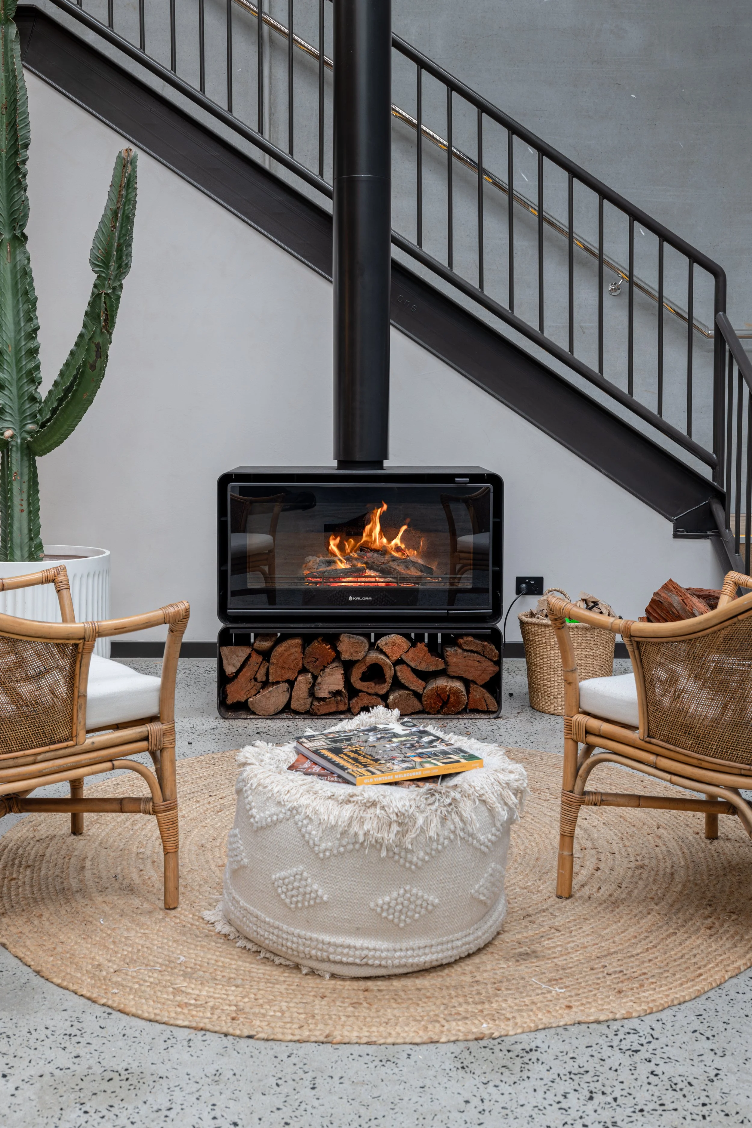 Modern indoor sitting area with two wicker chairs, a round woven rug, a cozy knit ottoman with magazines, a wood-burning fireplace with logs below, a large green cactus to the left, a black metal staircase with banister to the right, and gray concrete walls.