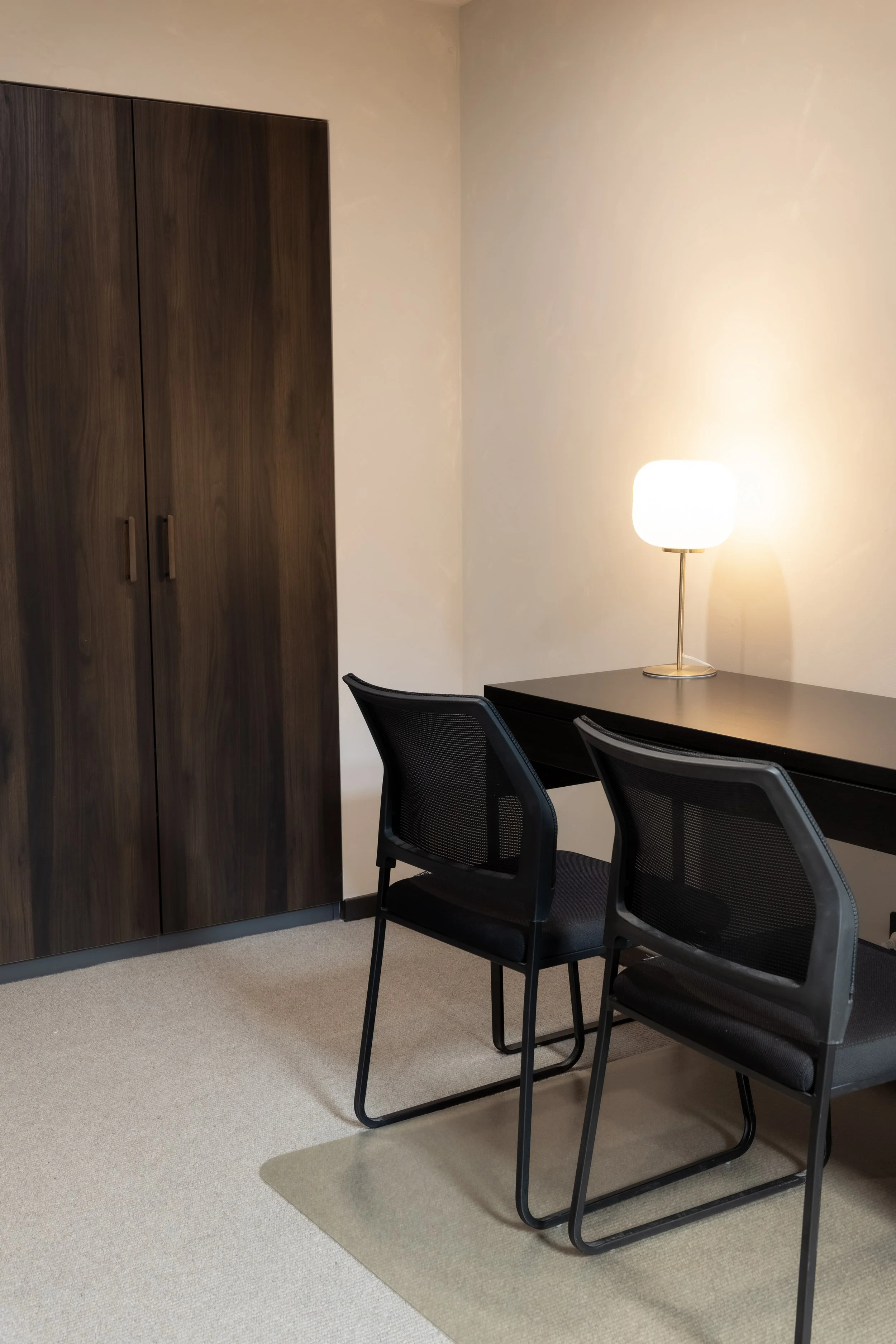 Office room with a dark wooden cabinet, a black table, black chairs, beige carpet, and a white lamp on the table illuminating the area.