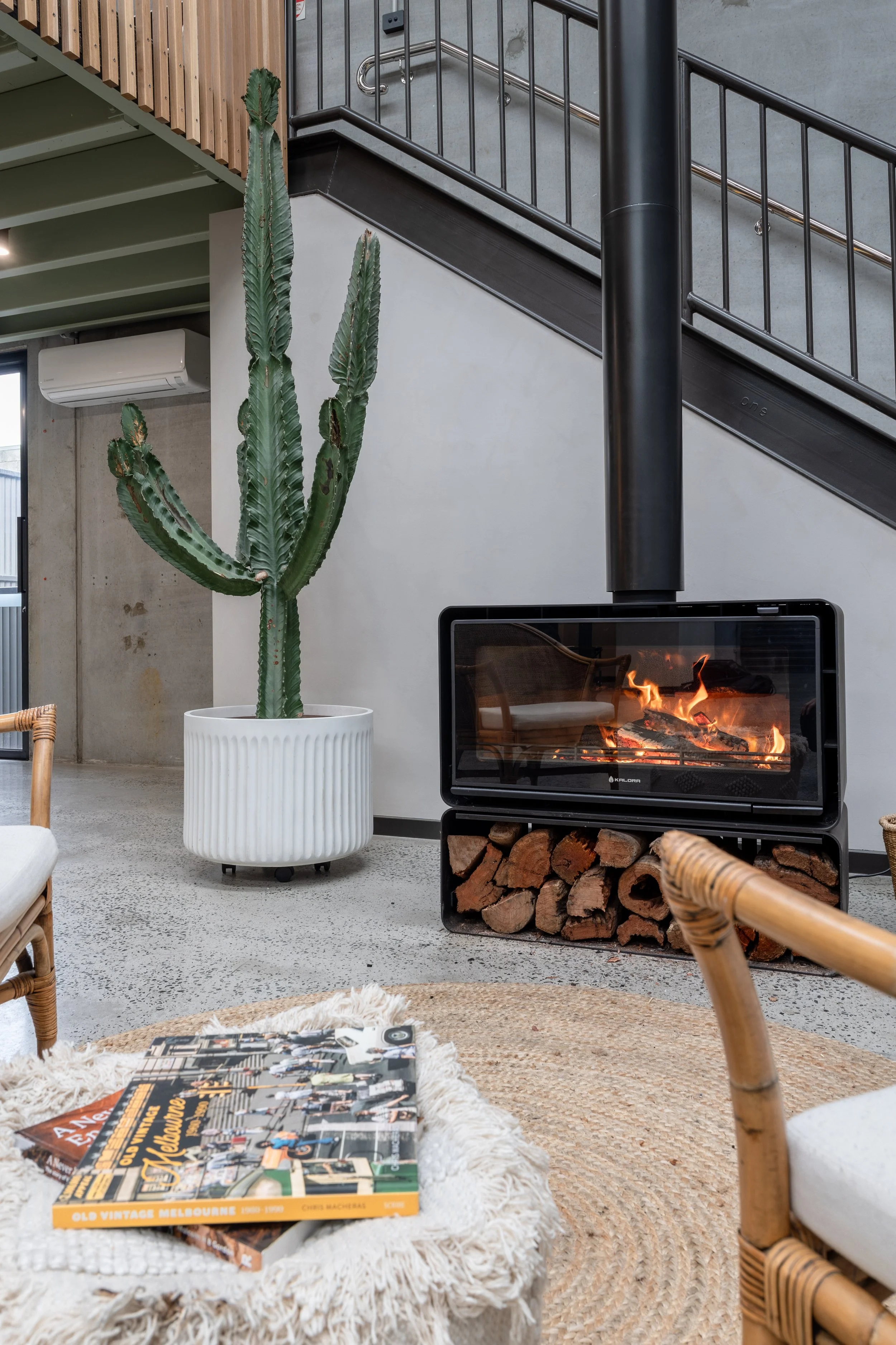 Indoor modern living space with a tall cactus in a white pot, a fireplace with fire, logs underneath, a woven rug, a magazine on a white fuzzy blanket, wooden furniture, a staircase with metal handrails, a wall-mounted air conditioner, and contemporary decor.