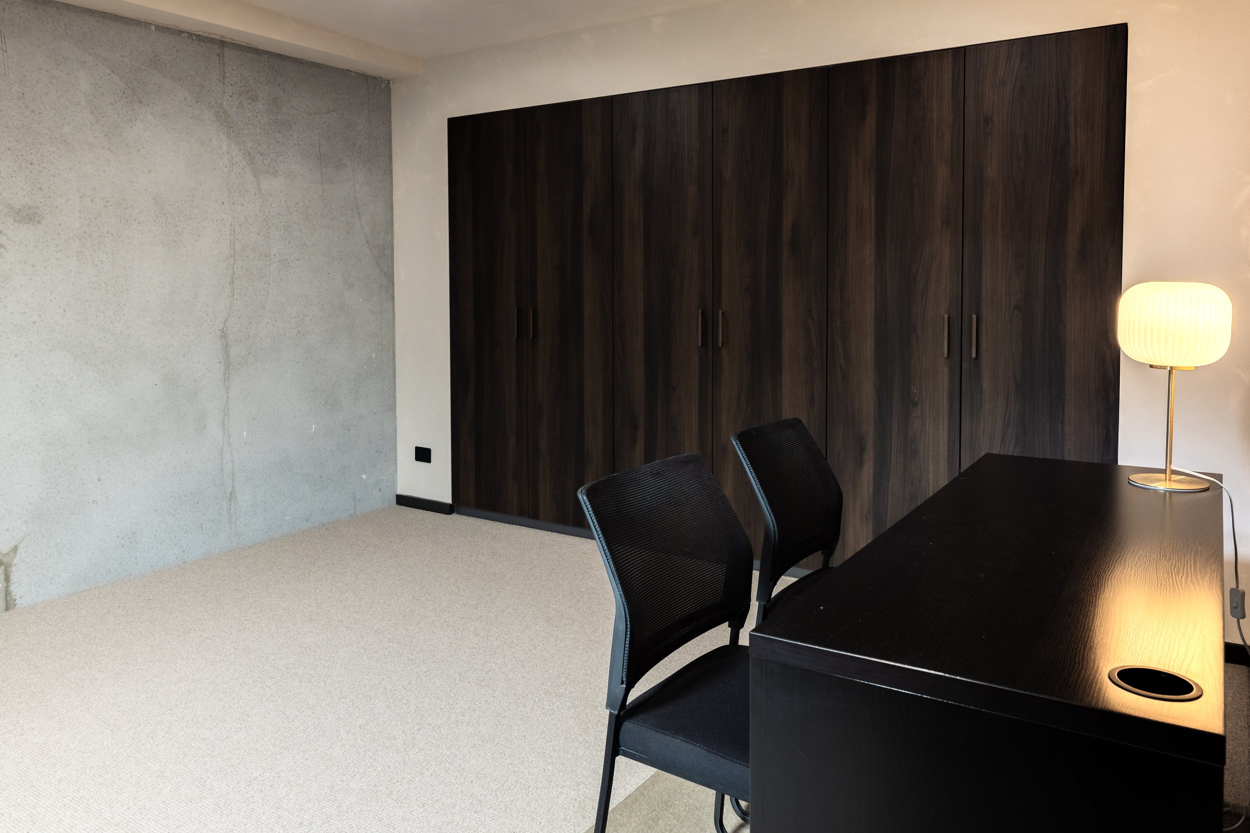 An office room with a dark wooden cabinet, a black table, black chairs, a desk lamp with a white shade, and a concrete wall, with a beige carpeted floor.