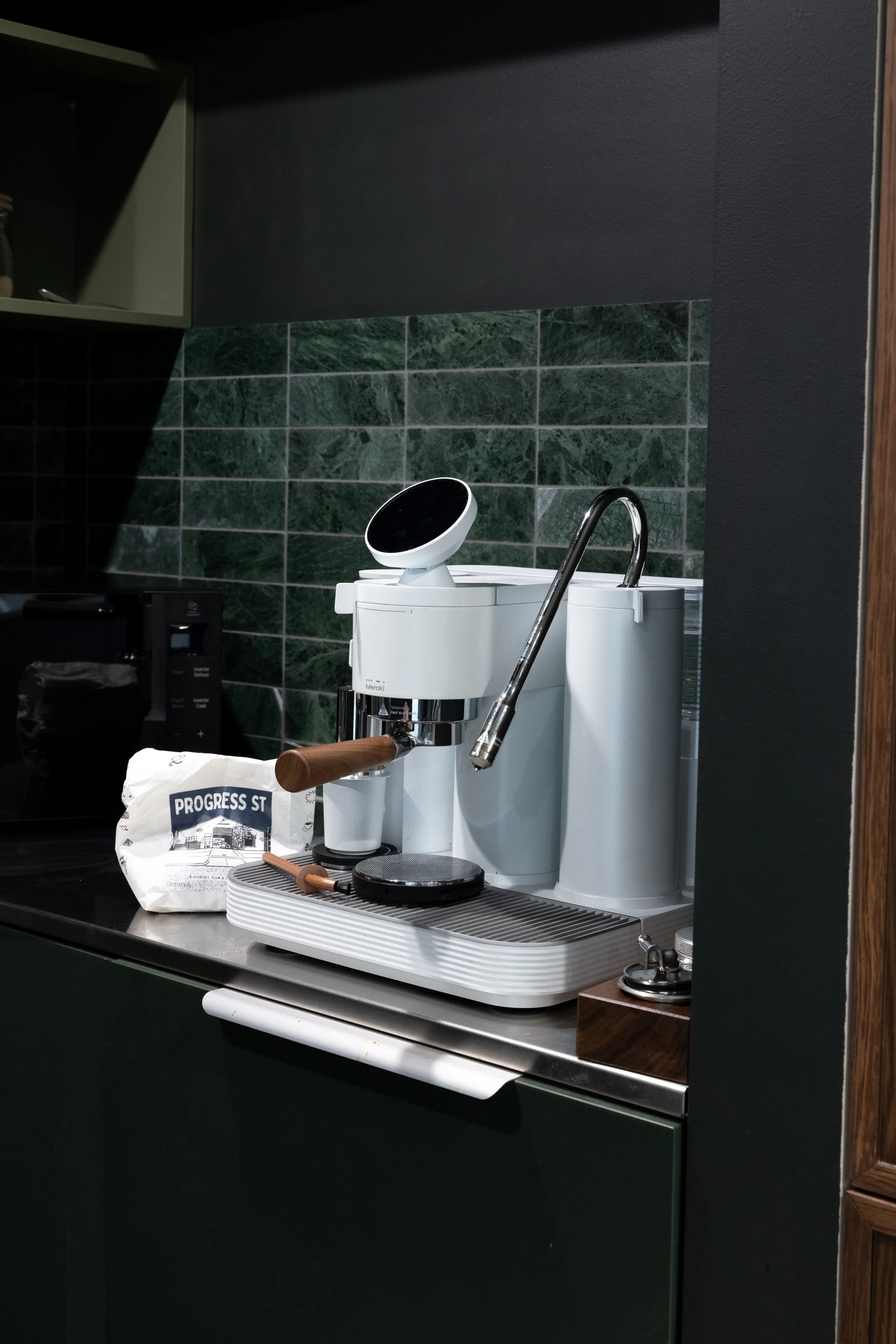 A coffee station with a white espresso machine on a countertop, a bag of coffee beans labeled 'Progress St,' a coffee tamper, and a coffee scale, set against a green marble tile backsplash.
