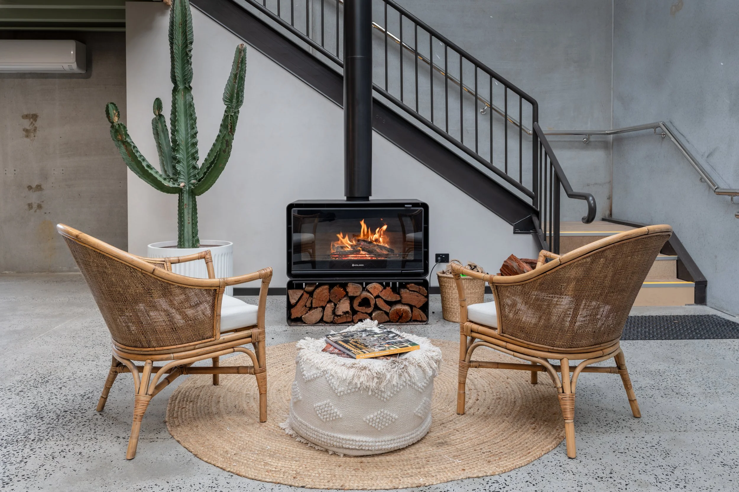 Modern indoor living space with two wicker chairs around a small round woven ottoman with a fluffy cream-colored blanket, a rectangular wood fire heater with a visible fire, a large potted cactus, and a staircase with black metal railing against a gray concrete wall.