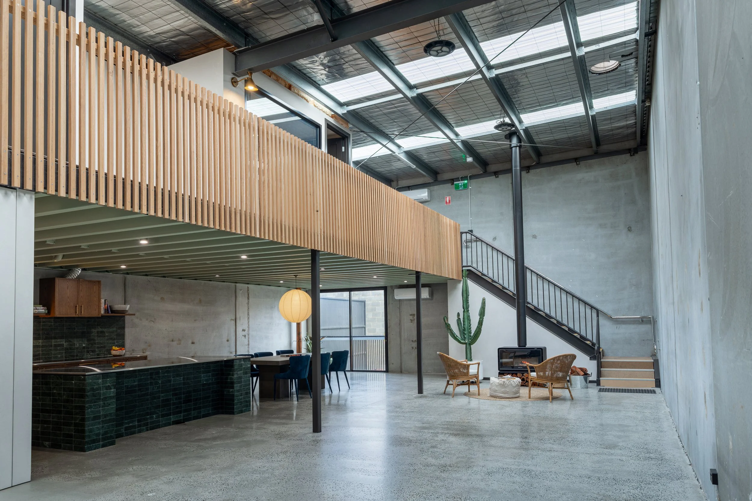 Interior of a modern industrial-style building featuring a high ceiling with exposed metal beams and skylights, a wooden slat balcony, a staircase with black railings, green tiled bar area, and minimalist furniture including wicker chairs, a cactus plant, and a hanging paper lantern.