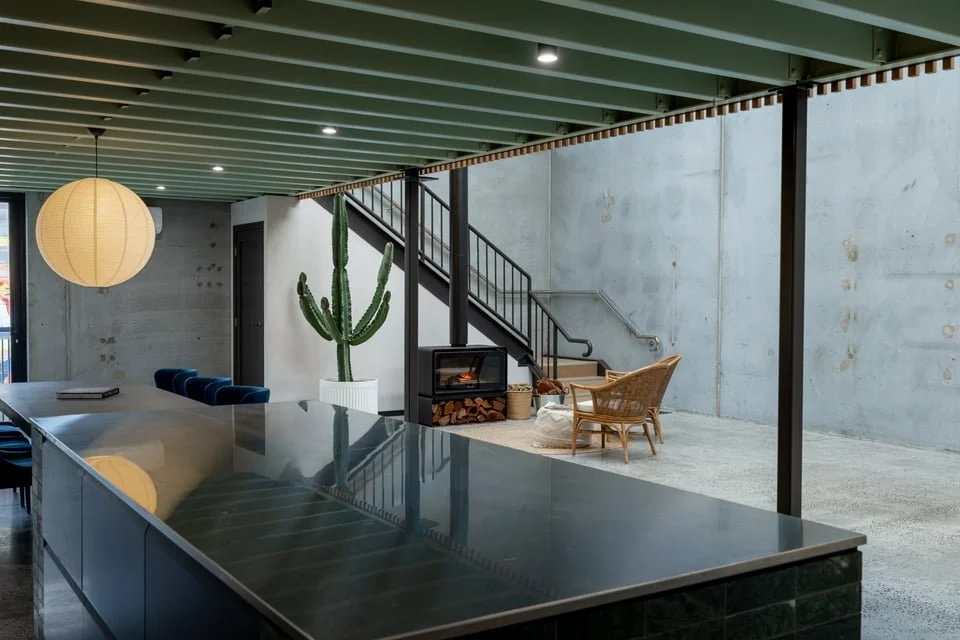 Modern interior space with a high ceiling, concrete walls, a large counter with a reflective surface in the foreground, a hanging paper lantern, a tall cactus plant, a wood stove with stacked firewood, rattan chairs, and a staircase.