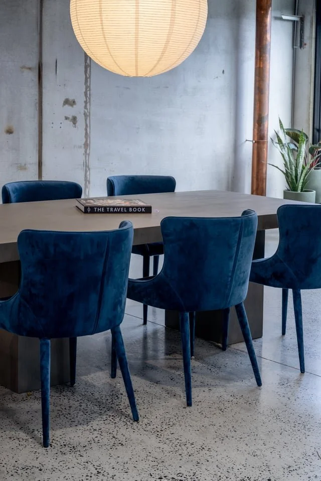 Modern dining room with a concrete wall, a large paper lantern, a concrete top table, and six blue velvet chairs. A travel book is on the table, and a potted plant is in the corner.