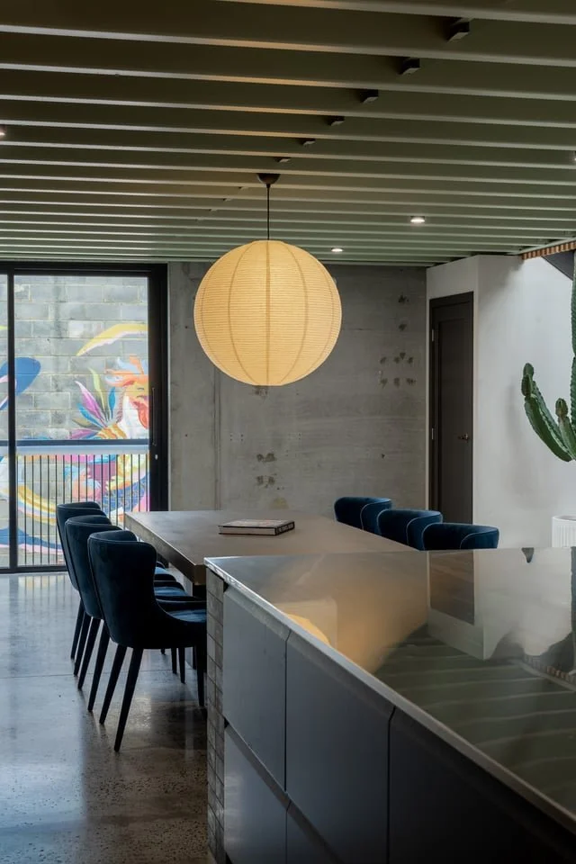 Modern dining area with a large white dining table, six dark blue chairs, a hanging paper lantern light, a large window with a colorful mural outside, and a door on the right side.