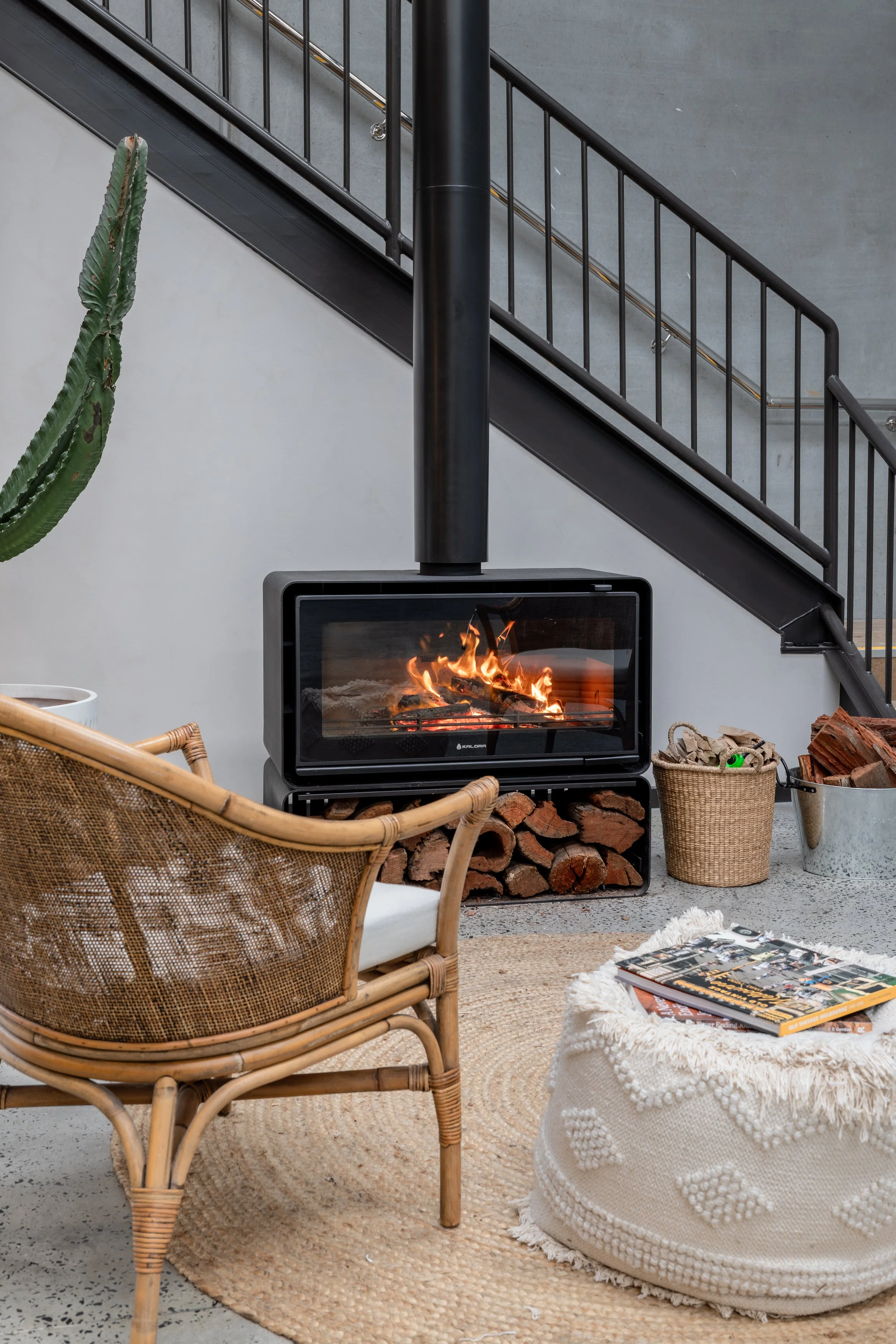Indoor space featuring a fireplace with visible flames, a wicker chair, a woven rug, a pile of firewood, magazines on a knit pouf, a cactus plant, and a staircase with black metal railings and concrete wall in the background.