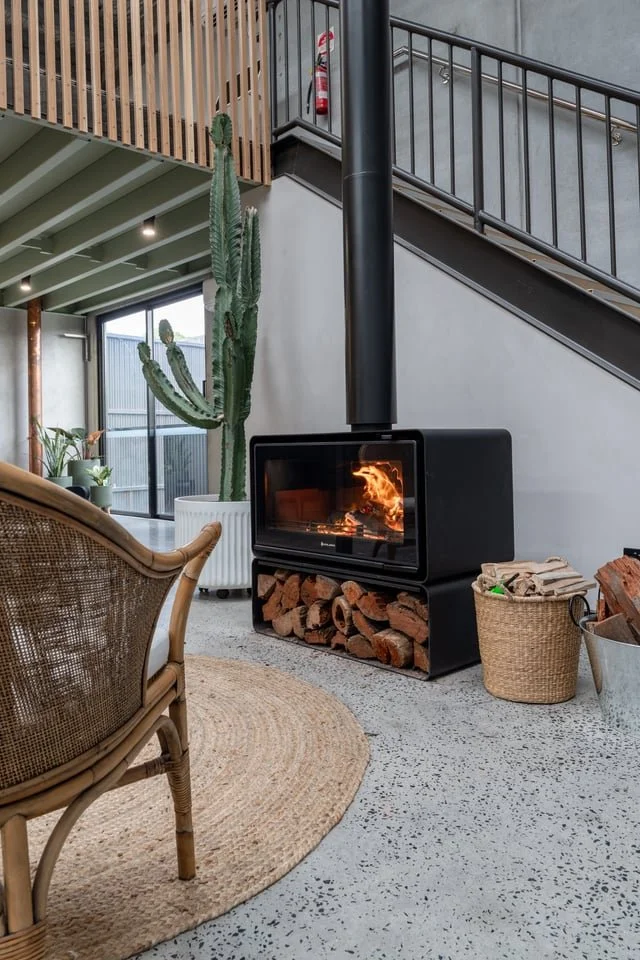 Modern living room with a wood-burning stove, a tall cactus in a white pot, rattan furniture, and baskets with firewood.