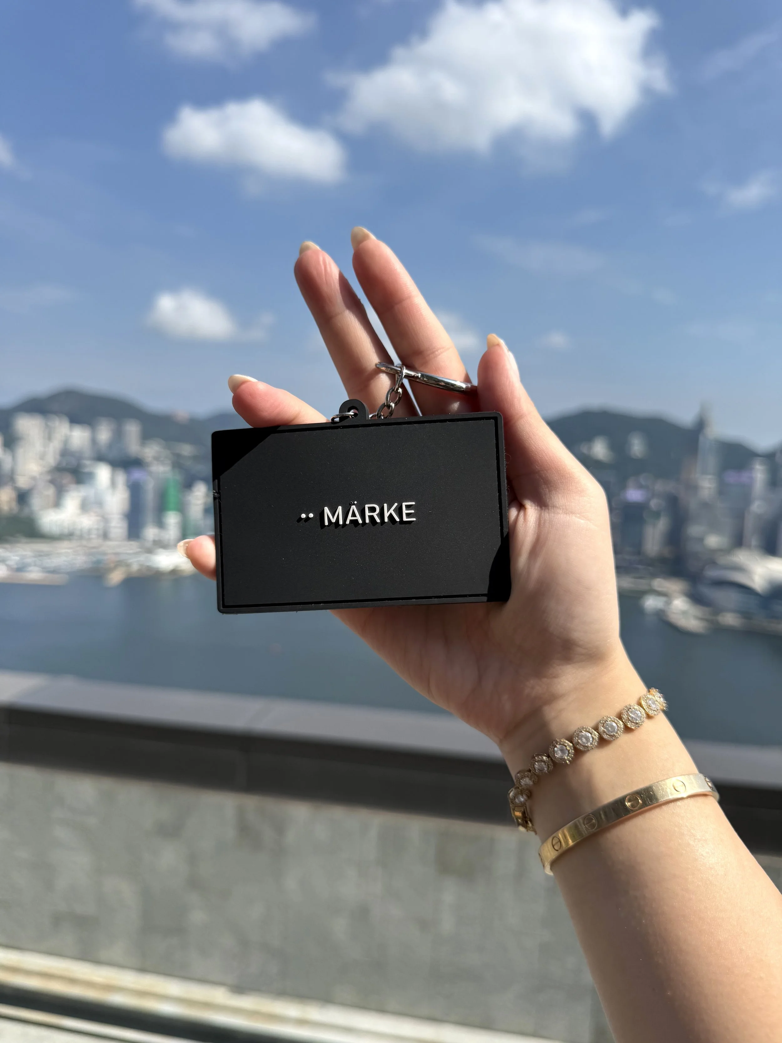 A person's hand holding a black object with text and a chain, against a cityscape with buildings, mountains, and a blue sky with clouds in the background.