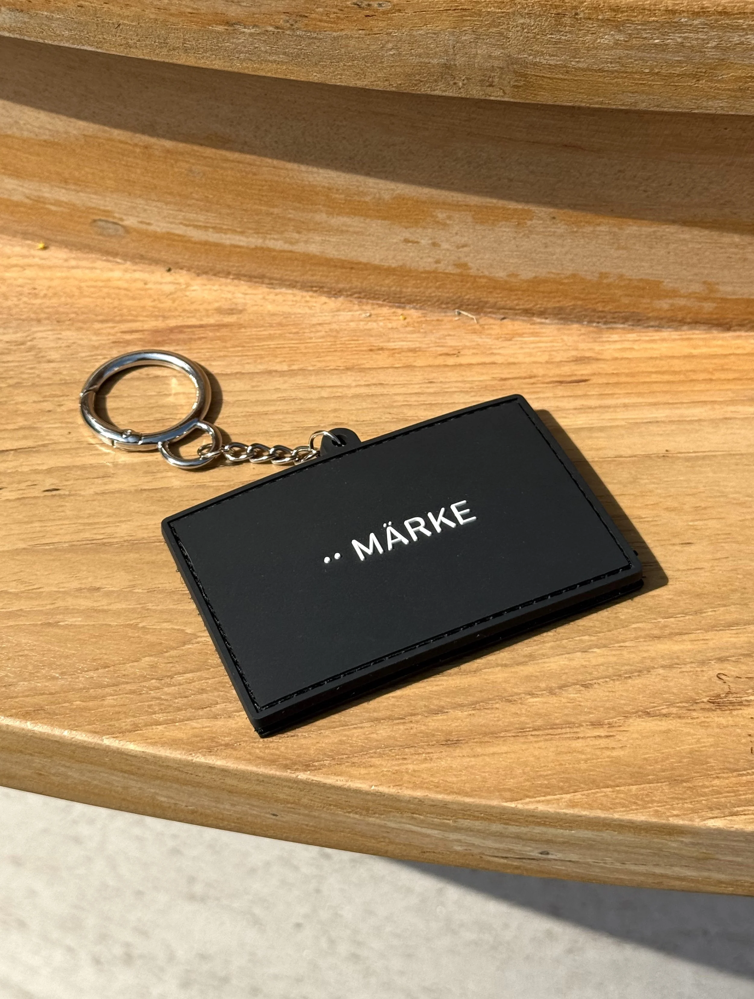 Close-up of a black leather keychain with the word '..MÄRKE' in white, attached to a silver key ring on a wooden surface.