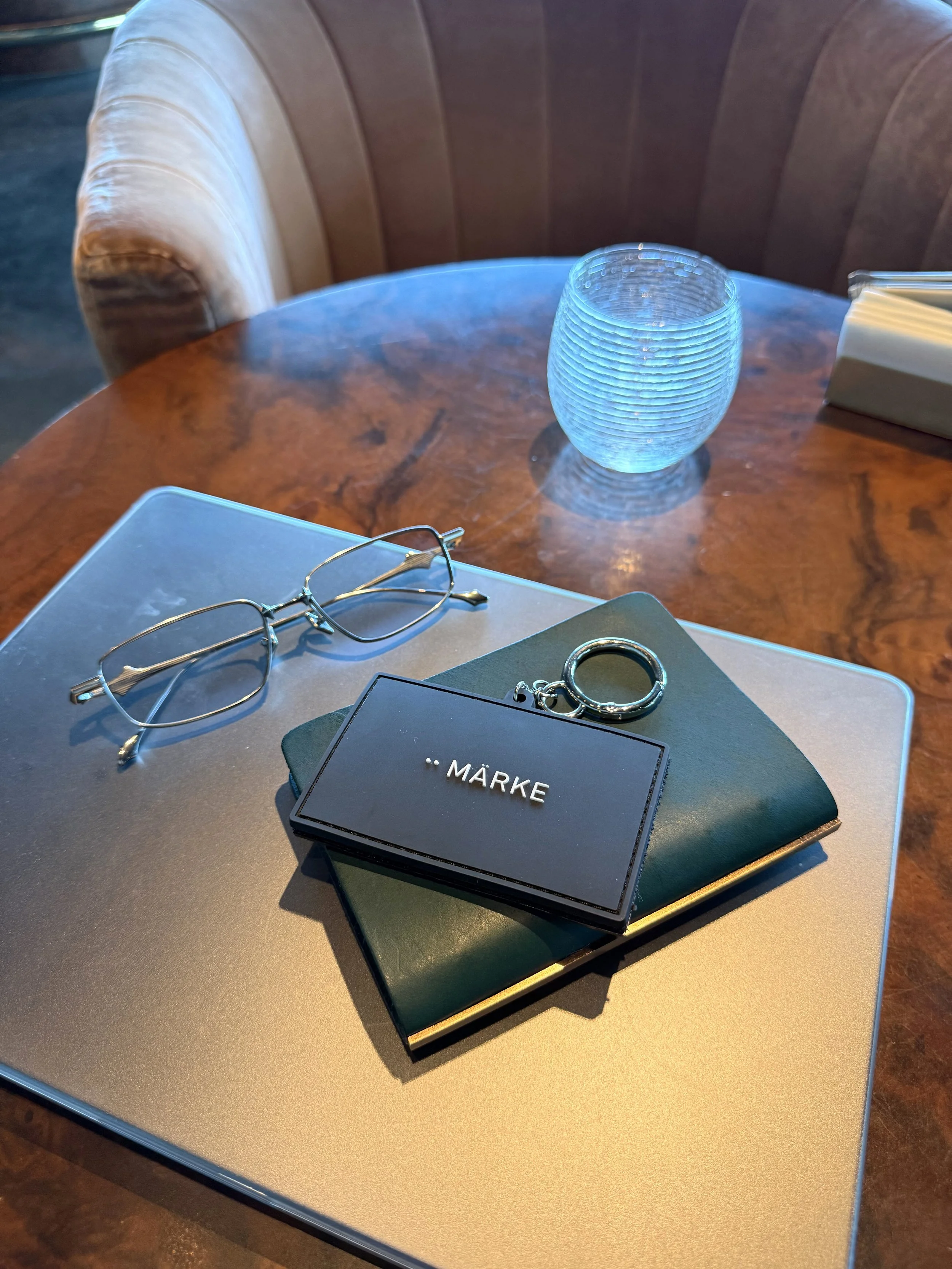 A round wooden table with a glass vase, a pair of eyeglasses, a closed black box with the word 'MÄRKE' on it, a closed laptop, and a white tissue box with pencils on top. In the background, there is a section of a brown leather booth.