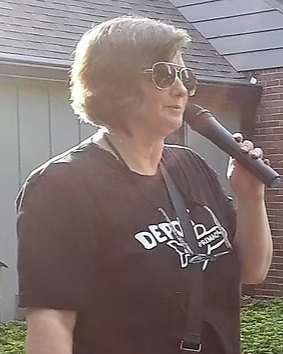 Older woman with short gray hair and sunglasses holding a microphone, standing outdoors in front of a house.