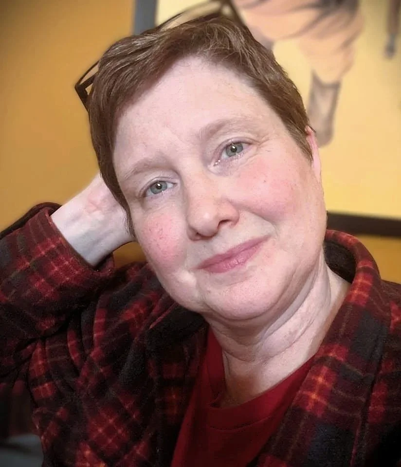 A close-up of Ames, a non-binary person, with short red hair, wearing a red and black plaid shirt, smiling gently at the camera, sitting in front of a yellow wall with a piece of artwork.