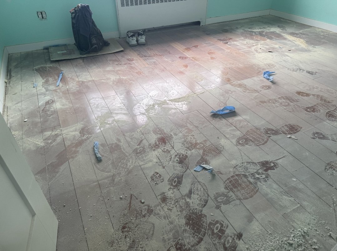 Room with partially installed flooring, scattered blue painter's tape, footprints, and dust. A backpack and shoes are in the corner.