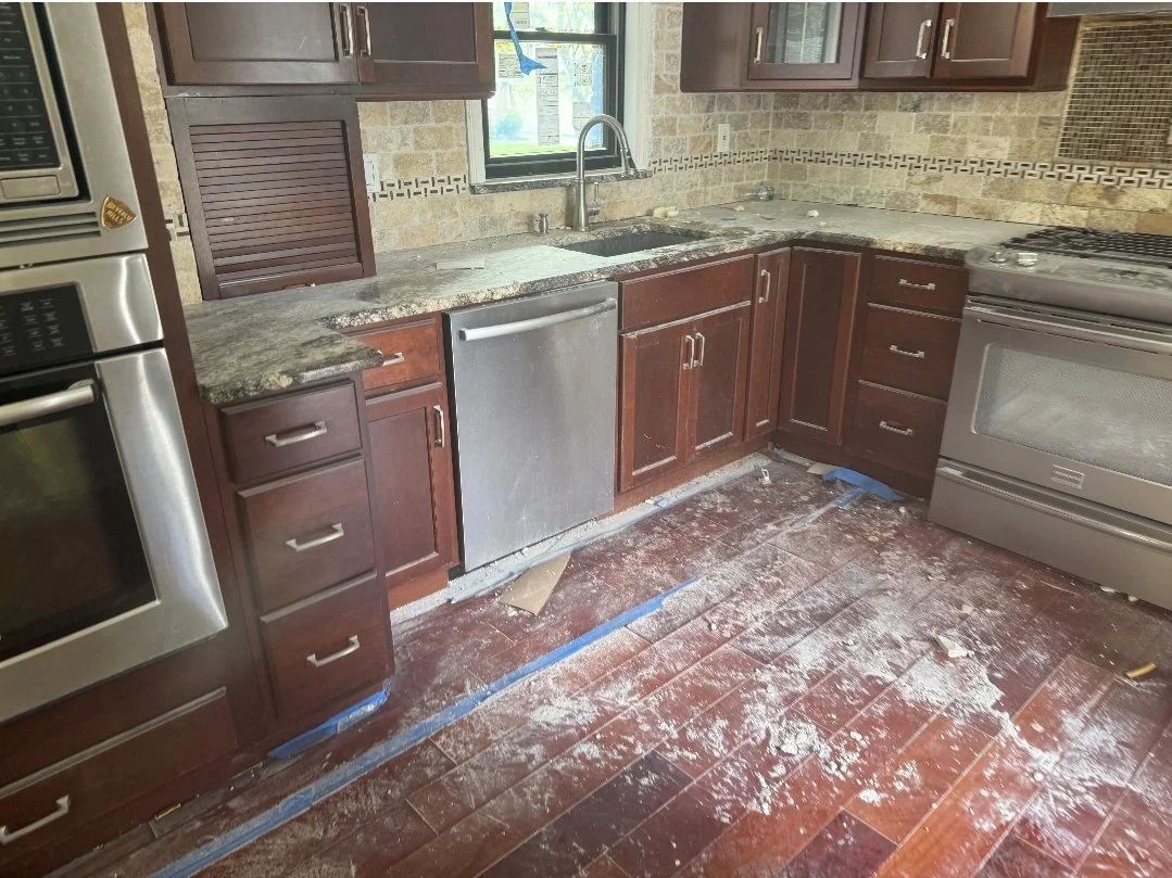 A kitchen under renovation with dust and debris on the wooden floor, dark wood cabinets, a granite countertop, a stainless steel dishwasher, and a stove, with a window above the sink.