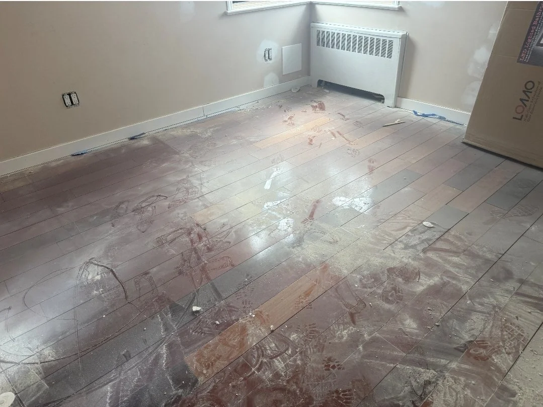 Room with unfinished wooden floor, covered in dust, footprints, and construction marks. The wall has loose electrical outlets and a radiator below the window. A large cardboard box leans against the wall.