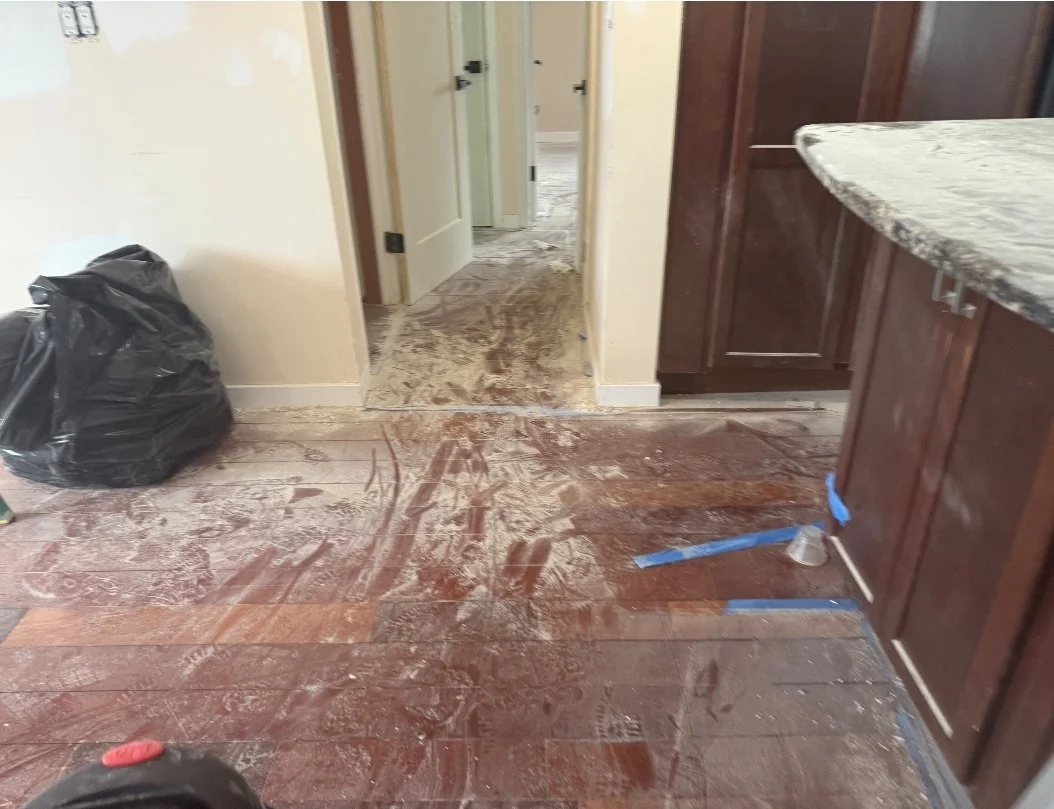 Floor in a room covered with dirt and debris after a renovation or cleanup, with a black trash bag on the left and blue painter's tape on the right.