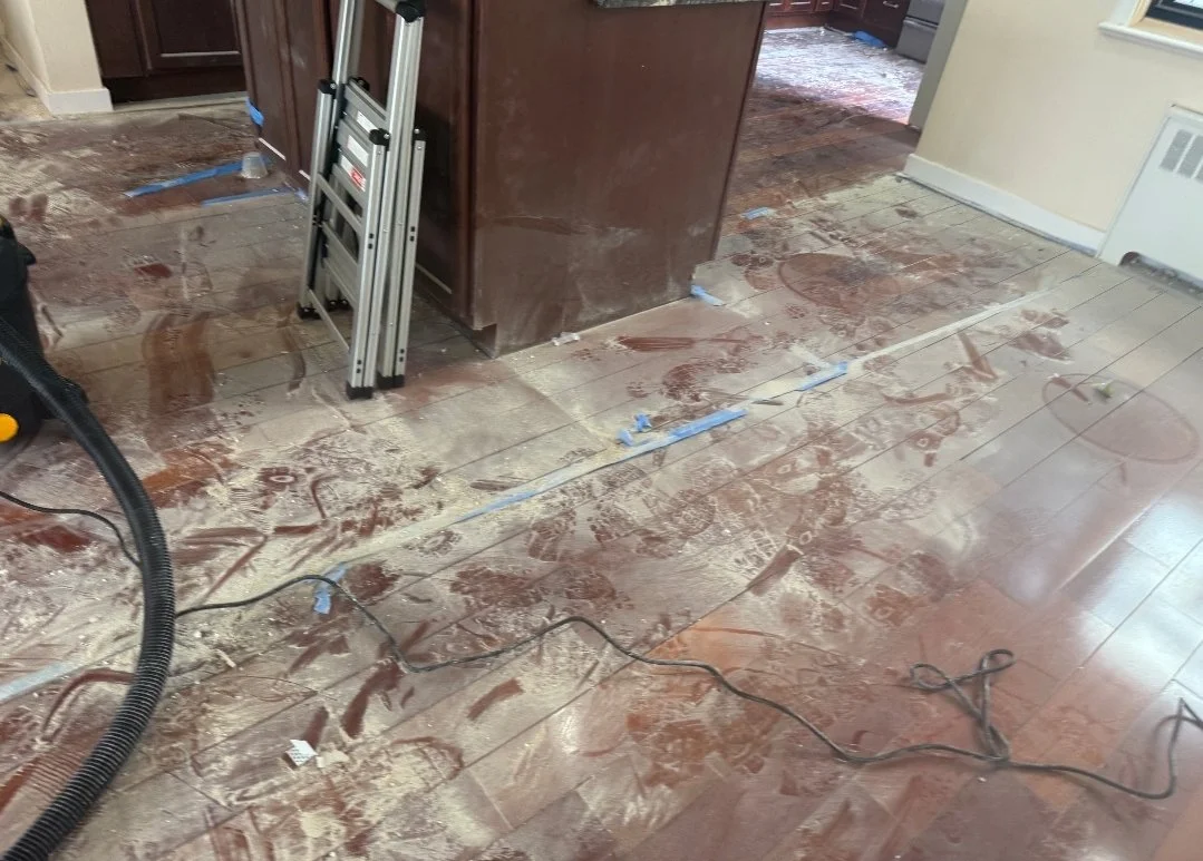 A kitchen floor undergoing renovation, with scattered debris, dust, and shoe prints. Construction tools and electrical cords are visible, along with a brown cabinet and a step ladder leaning against it.