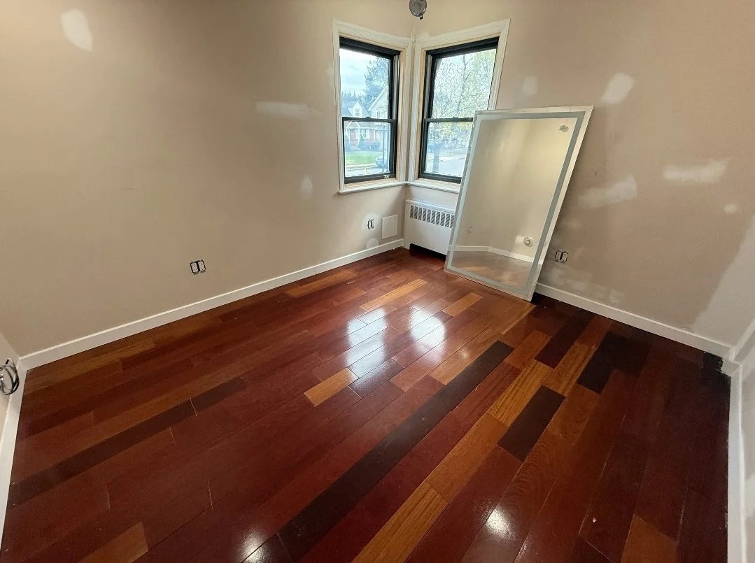 Empty room with beige walls, hardwood floor, two windows, and a large mirror leaning against the wall.