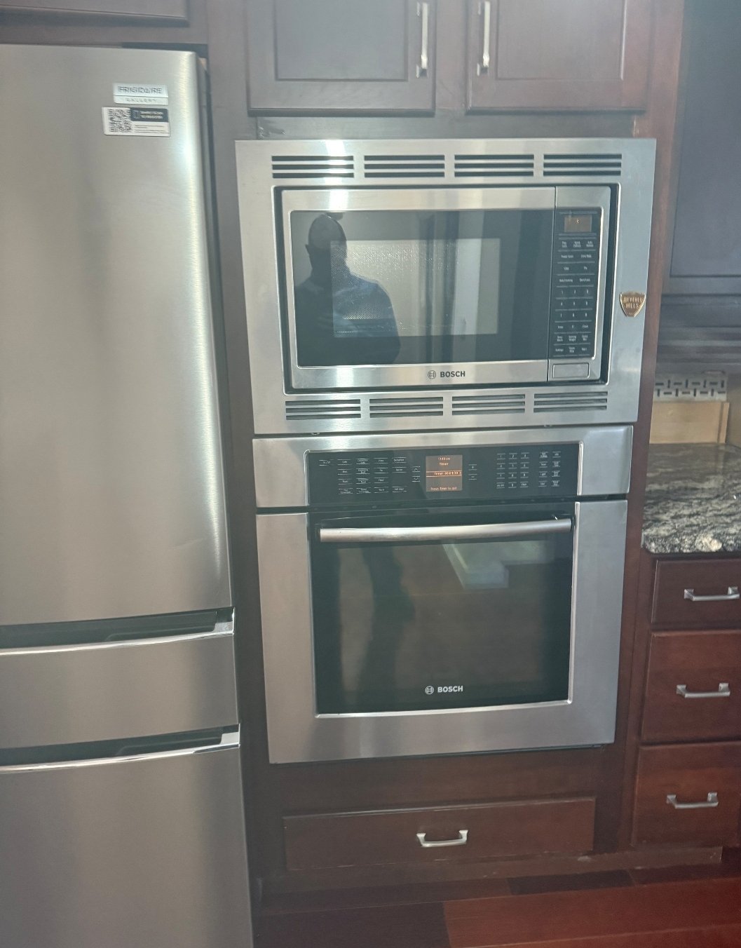 Kitchen with a stainless steel microwave and oven by Bosch, built into wooden cabinetry, next to a refrigerator and granite countertop.