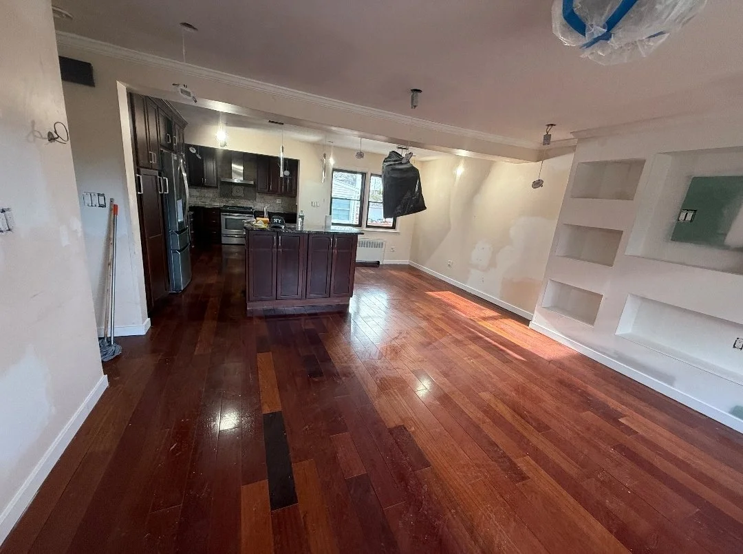 An empty kitchen and living area with hardwood floors, kitchen cabinets, and a window. There is a black plastic bag hanging from the ceiling, unfinished walls, and some construction materials.