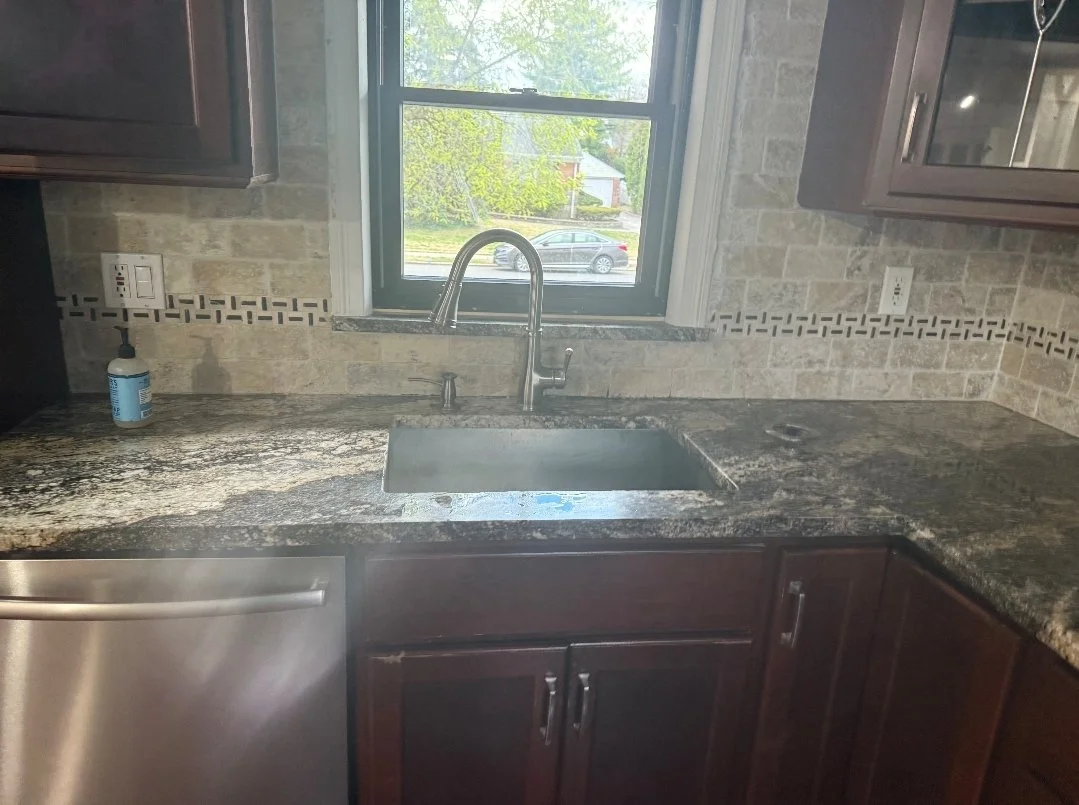 Kitchen countertop with a stainless steel sink, faucet, dark wooden cabinets, a bottle of soap, and a window showing a street and trees outside.