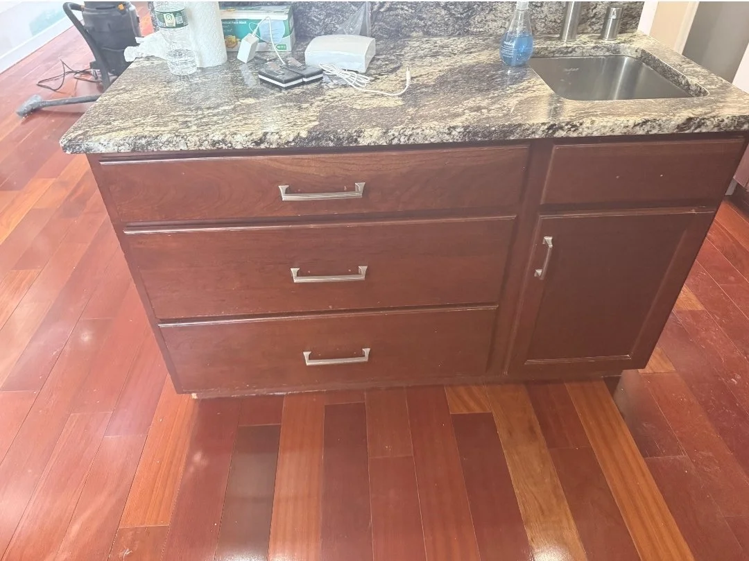 Kitchen island with dark granite countertop, wooden drawers with metal handles, and a small sink with a soap dispenser nearby.