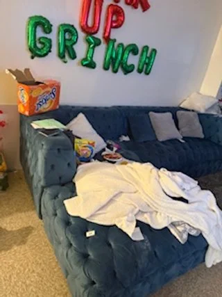A blue sofa with a white cloth draped over it, a box of snack crackers on the armrest, and a pile of clothes or linens on top. The wall behind has colorful balloon letters spelling "Wake Up Grinch."
