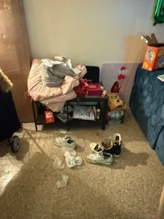Cluttered bedroom corner with shoes and trash on beige carpet, bed partially visible with blankets and pillow, small black table with items on it, and wall with a green light strip.