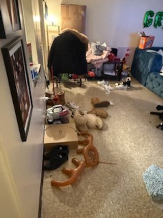 A cluttered living room with various toys, shoes, and clothing scattered across the floor, a person bending over near a small table, and a blue couch against the wall.