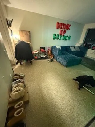 Living room with a blue sectional couch, a person sitting at a desk, and a sign on the wall that reads 'Dropping Up Crates' in red and green letters. There are shoes and other items in the room.