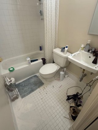 Small bathroom with a bathtub, toilet, and sink. Items like bottles, a laundry basket, and slippers are visible.