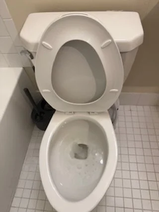 Toilet with the lid open, in a bathroom with white tiled floor and beige wall.
