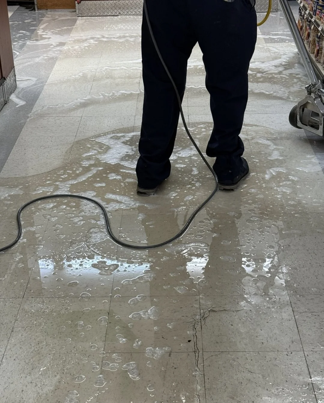 Person cleaning a wet, soapy floor with a floor scrubber in a store aisle.